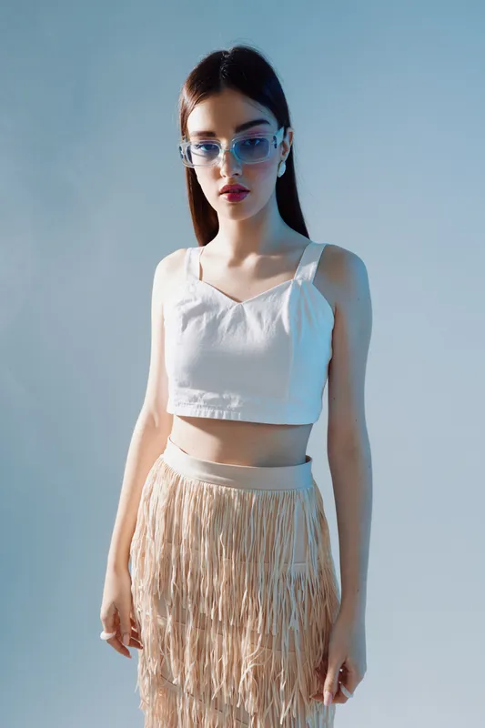 Young woman in stylish white crop top and fringed skirt poses confidently against blue background wearing translucent blue sunglasses. Concept for woman beauty fashion and modern style.