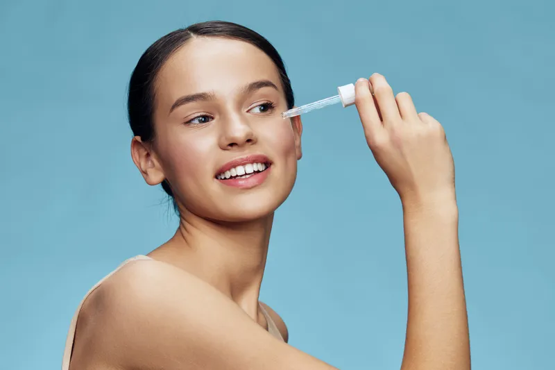 Young woman applying skincare serum for radiant skin on a blue background, showcasing healthy beauty and self care