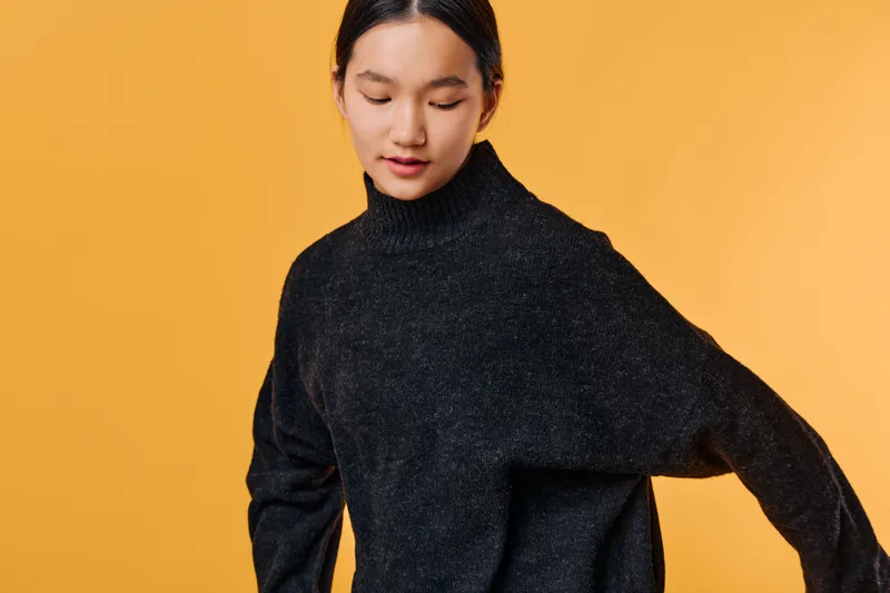 Woman with black sweater looking down posing on orange background casual style and minimal studio portrait with fashion concept and cozy clothing on model.