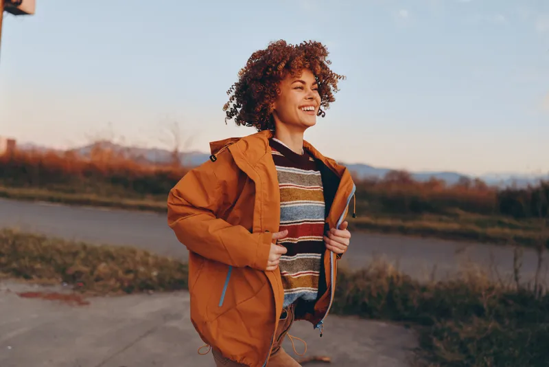 Woman lifestyle outdoor scene at sunset, golden hour, curly haired traveler in brown jacket carries backpack and walks along path by the lake, evening light, relaxed travel mood in nature