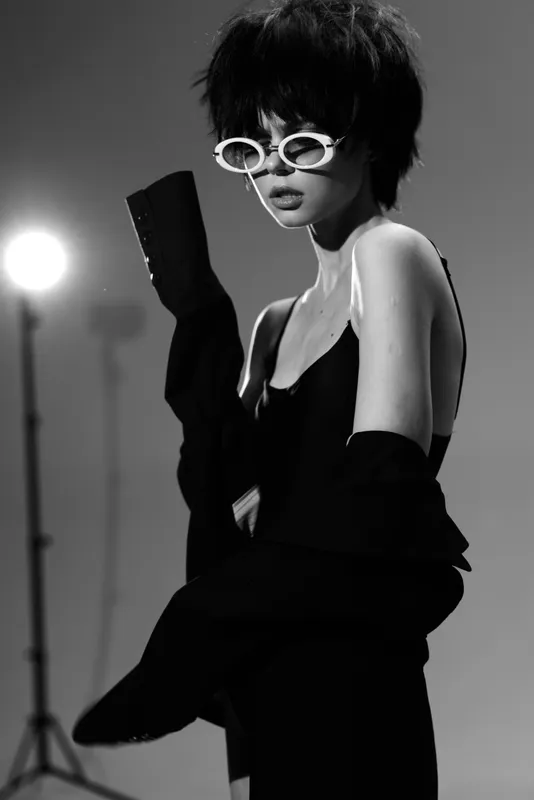 Stylish woman in white sunglasses and black outfit posing confidently in black and white studio setting