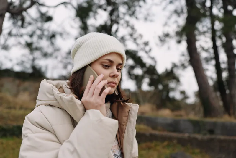 Woman outdoors in a coat and beanie on a cold winter day, talking on a phone, jacket visible, casual attire, calm portrait in a park with trees.