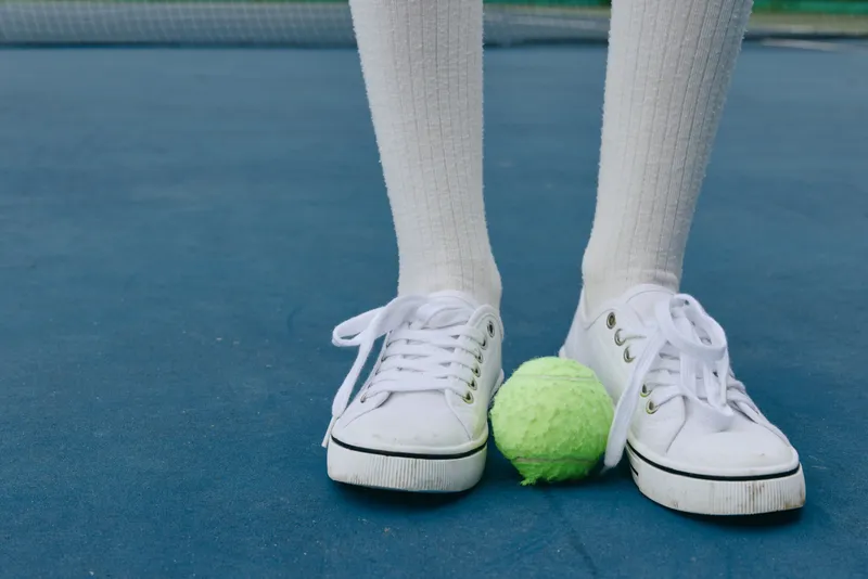 Young athlete wearing white sneakers and socks standing on a tennis court, focusing on a bright green tennis ball, symbolizing energy and sportiness