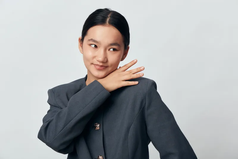 Woman in dark blazer smiling and posing with hand on shoulder against light background. Portrait of young female with neat hair looking sideways with calm expression.