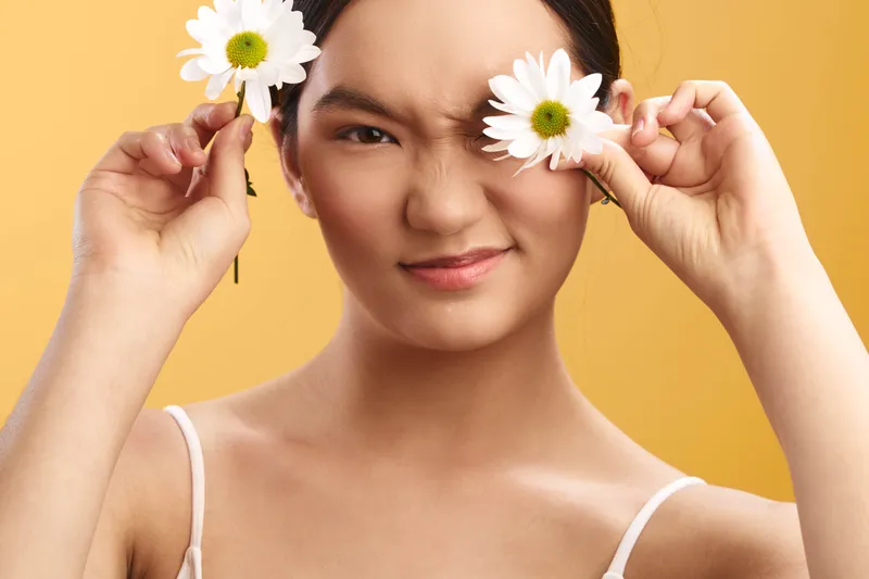 Cheerful asian woman holds white daisies near her eyes against a warm yellow background, creating a fresh, playful mood suitable for lifestyle, beauty and wellness projects.