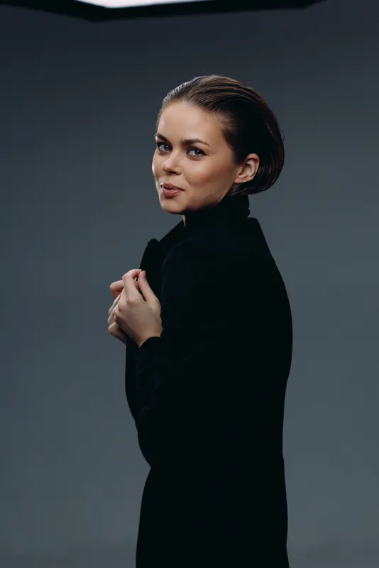confident woman in black coat posing with slight smile against dark background