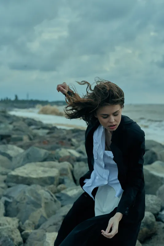 Woman in blazer and white shirt on a windy rocky beach, dramatic fashion scene by the sea, movement and moody atmosphere, outdoor shoot