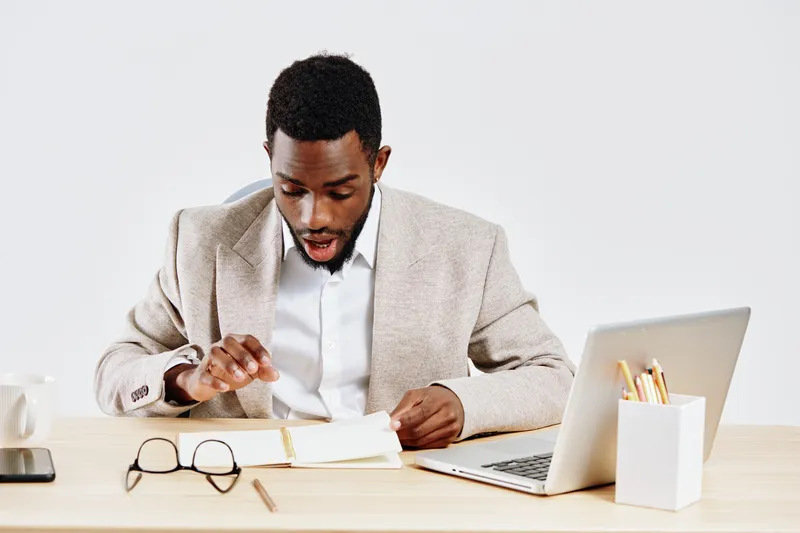Professional young African American man wearing beige blazer and white shirt working at desk with laptop, notebook, glasses, and coffee mug in bright modern office environment. Business and office con