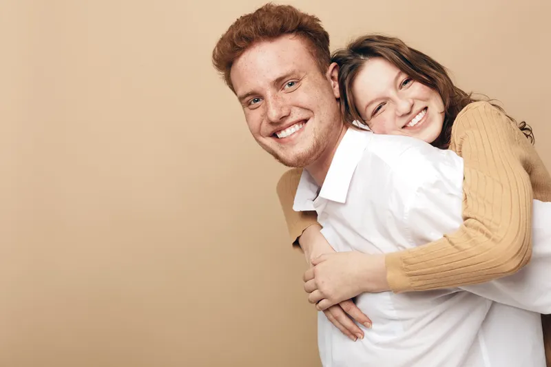 Happy young couple with red hair and brown hair smiling, woman hugging man from behind. Warm beige background, casual clothing, concept of love, friendship, and togetherness.