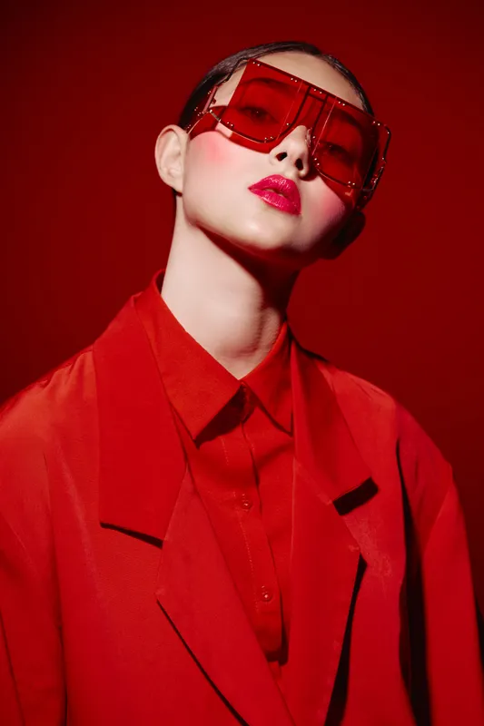 Fashion model wearing oversized red sunglasses and vivid red outfit poses against red background. Bold makeup with rose lipstick and stylized creative beauty concept for editorial work.