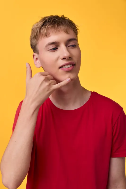 Young man in a red shirt making a friendly shaka gesture against a bright yellow backdrop, capturing casual style, confidence, and playful communication for lifestyle and fashion use