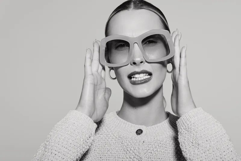 Stylish caucasian woman with pale skin wears oversized sunglasses and knitted sweater, posing on grey background.