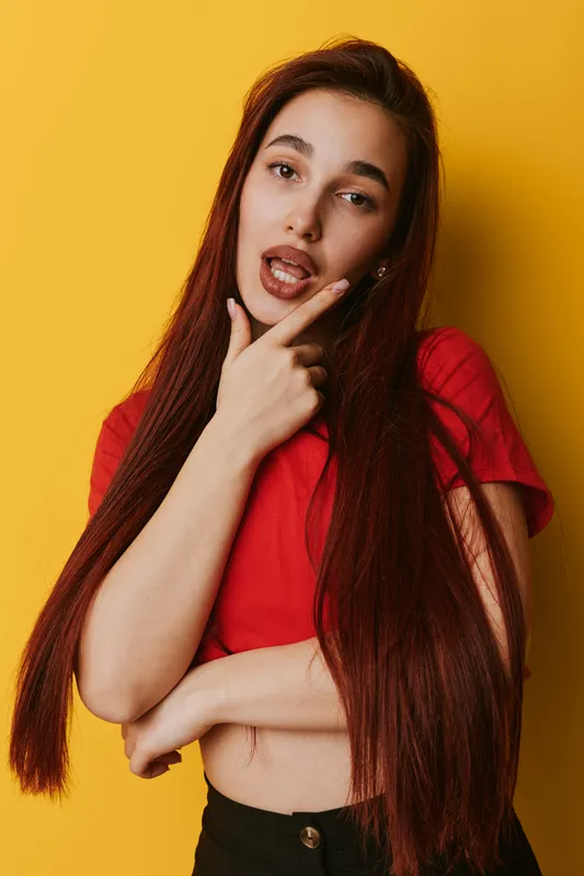 Young woman with long red hair wearing red shirt posing with thoughtful expression and hand on chin against yellow background in studio portrait of casual fashion.