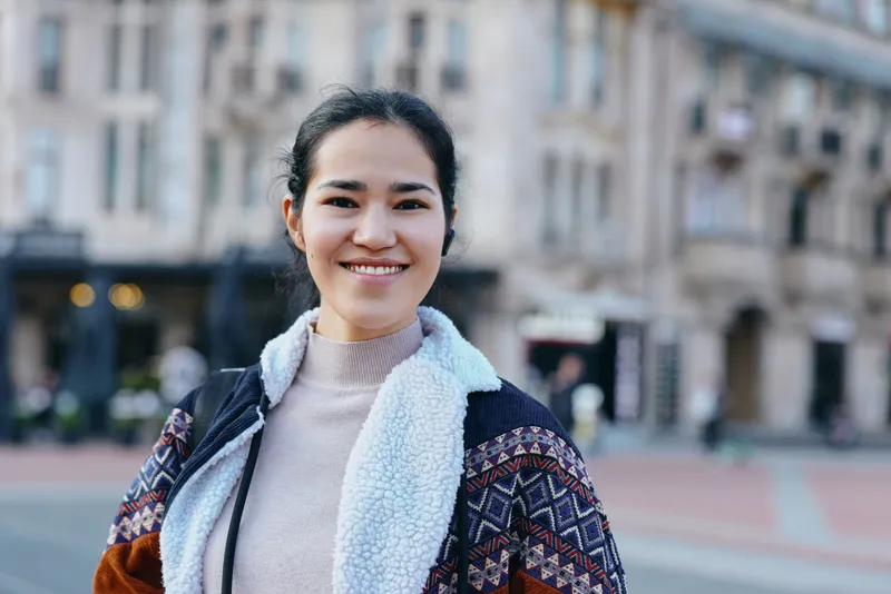 Smiling woman in a cozy sweater and scarf stands outdoors in a city street, casual fashion, warm light, urban backdrop, relaxed and confident, capturing everyday style and urban life.