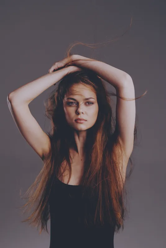 Portrait of a serious young woman with long hair and a black top posing with raised arms against a gray background.