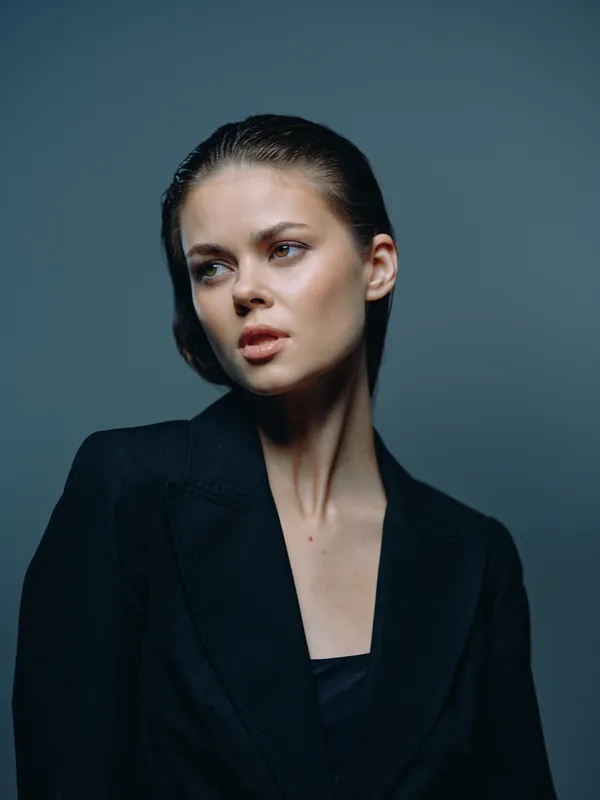 Portrait of a serious woman in black jacket with natural makeup and slicked-back hair, featuring a dark moody background and minimalist style.