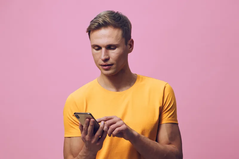 Smiling man using mobile phone in casual yellow t-shirt isolated on pink background studio portrait, people lifestyle concept, modern technology, communication, leisure, bright colors, cheerful mood