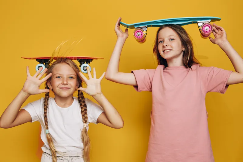 Two happy girls with skateboards pose against a yellow background. One girl holds a skateboard above her head while the other balances one on her own head. Concept for childhood and fun.