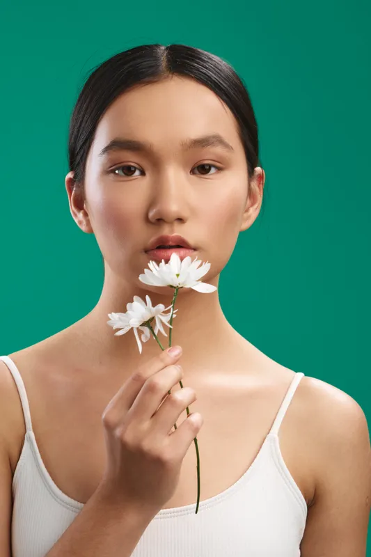 Asian woman gazes softly at the camera while holding a white flower, set against a vibrant green backdrop; a calm, minimalist portrait that conveys grace and focused presence.