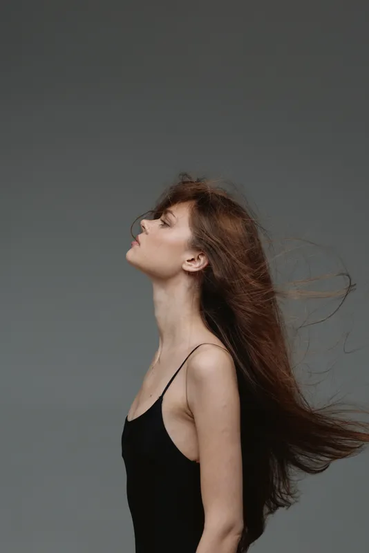 Profile of a young woman with long hair flowing, wearing a black dress, exuding calm and grace against a neutral gray background.