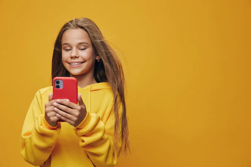 Girl in yellow hoodie using smartphone smiling and looking at screen standing against orange background. Young teenager with long hair enjoying mobile technology communication.