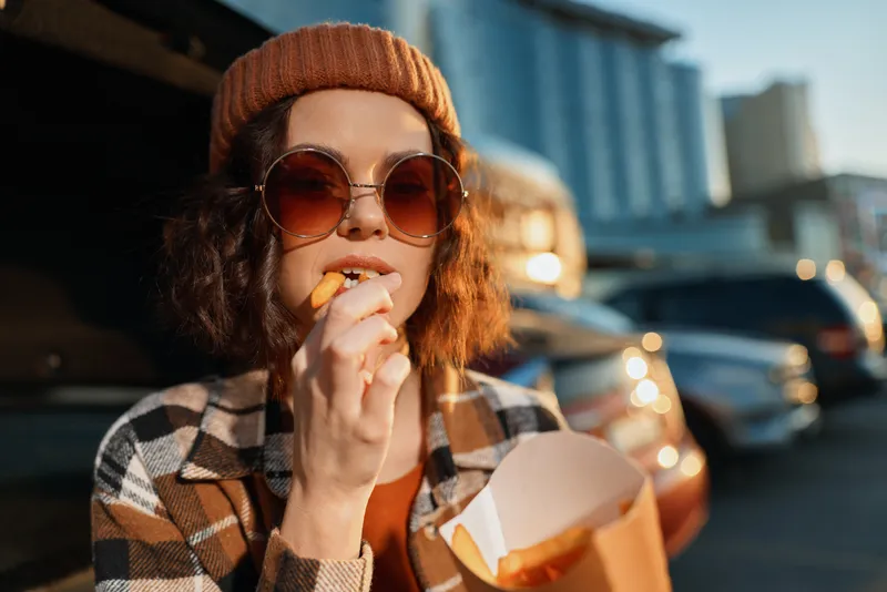 Woman eating fries from a paper container in a parking lot at golden hour, sunglasses and beanie, candid authenticity and emotional storytelling capturing mindful living urban lifestyle.