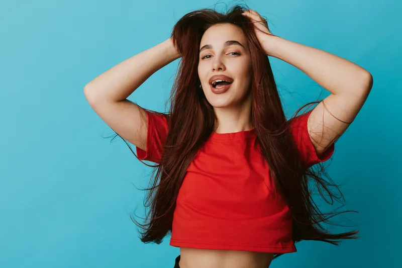 Young woman with long hair wearing red shirt posing with hands in hair smiling with closed eyes on blue background. Casual style, beauty and happiness concept portrait.