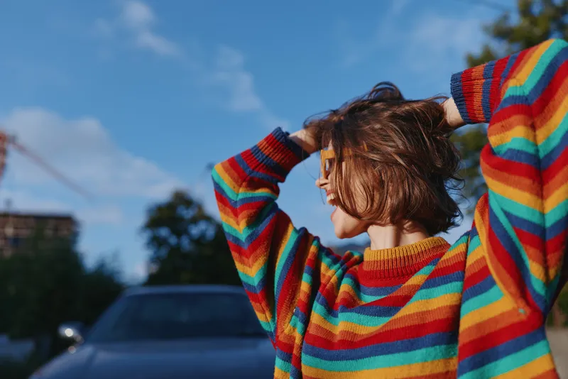 Woman in rainbow sweater with short hair smiling outdoors by a parked car, young adult leaning back with hands in hair, casual outfit in sunny urban lifestyle scene for travel and joy.