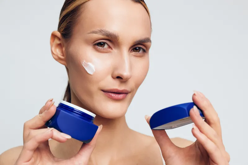 Young woman applying moisturizing cream on her face, holding blue jar and lid, clear skin, natural light, closeup portrait, skincare routine, beauty concept