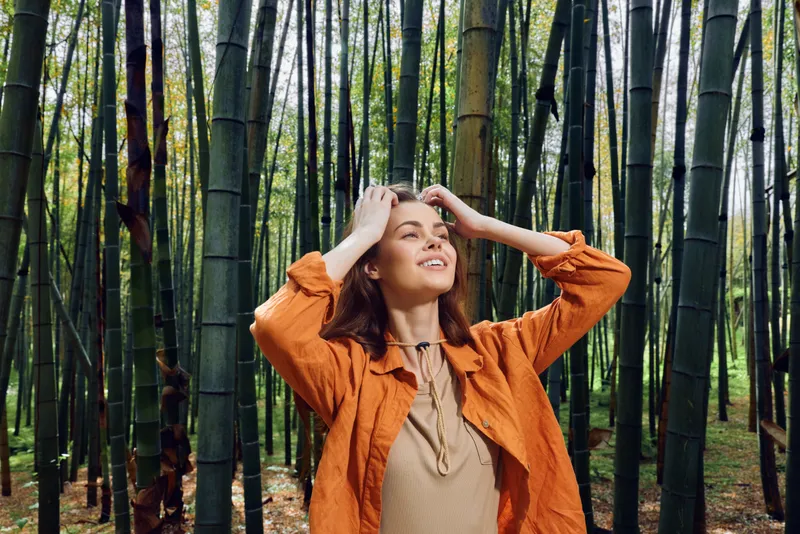Woman in bamboo forest smiling and looking up, portrait of a happy young female outdoors in orange jacket, nature, travel and mindfulness moment among tall green bamboo stalks.