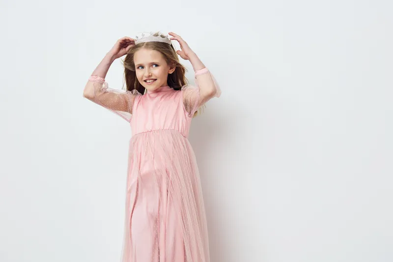Young girl in a pink dress with a floral crown, smiling joyfully against a light background, perfect for children s fashion and seasonal celebrations