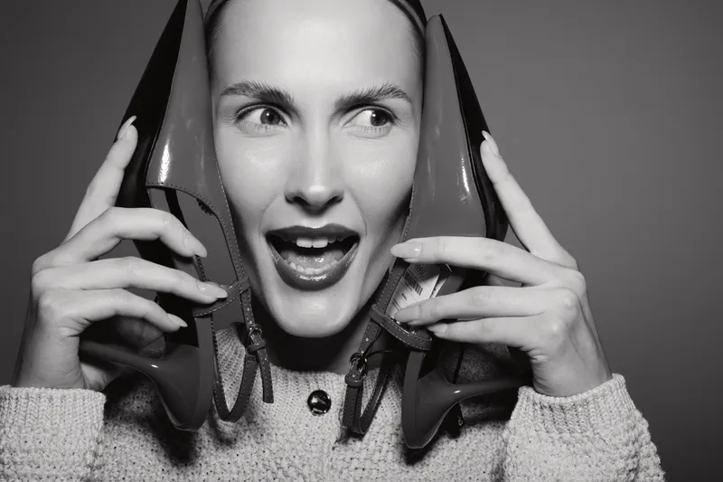 Caucasian woman with fair skin holds two high heel shoes near her face, expressing surprise and joy in studio lighting. Concept of fashion and emotions.