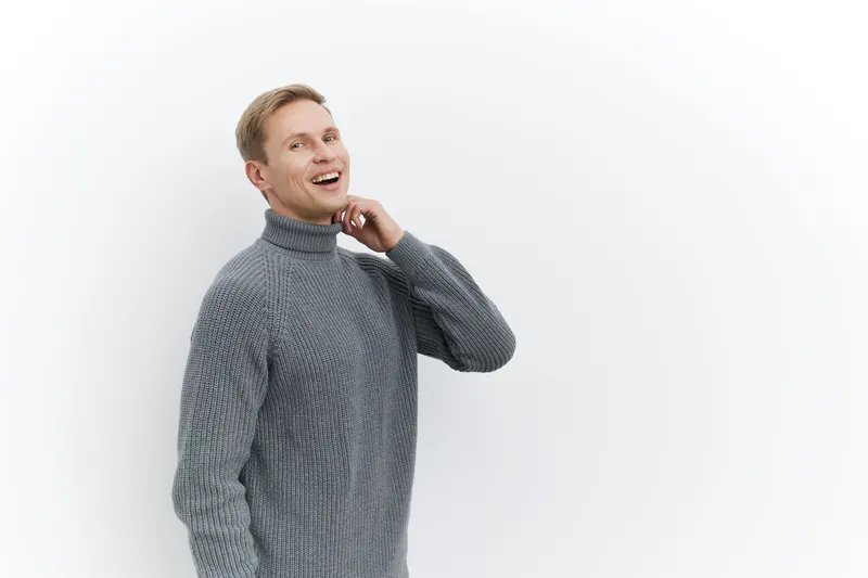 Smiling young man wearing a cozy gray sweater, looking cheerful and relaxed, standing against a plain white background in studio portrait. People's lifestyle concept.