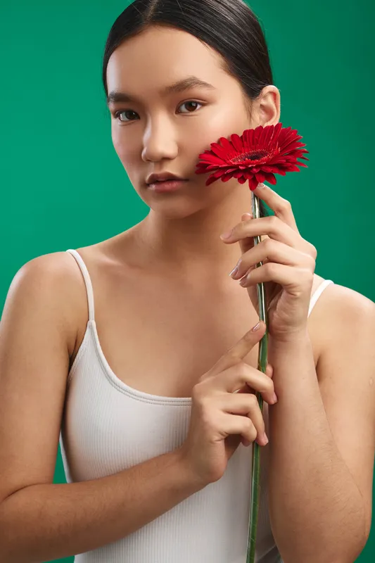 asian woman holding a red flower in a studio setting with a green backdrop. She gazes softly at the camera, creating a calm, elegant portrait that conveys serenity and focus.