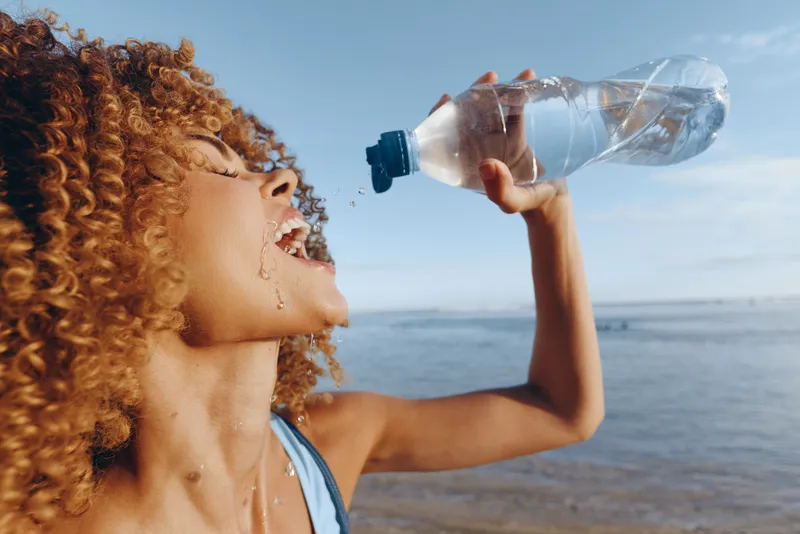 Woman drinking water from plastic bottle, curly hair profile, seaside outdoors in summer, hydration and refreshment concept, closeup with ocean horizon, natural sunlight, fresh breeze