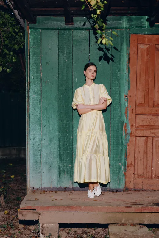 woman dress porch wooden standing portrait vintage authenticity inclusivity, young woman in yellow dress standing on rustic wooden porch by weathered green wall and closed door