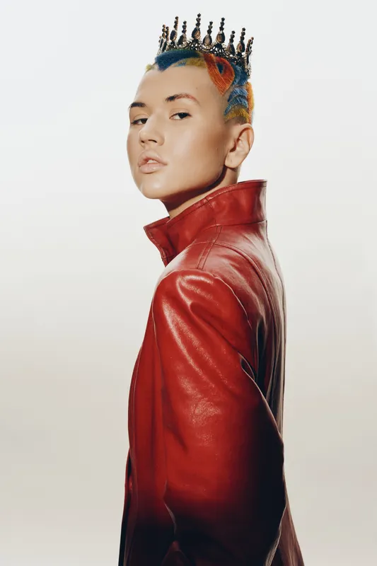 Young woman with colorful hair and a crown, wearing a red leather jacket, posing confidently against a bright neutral background in a modern fashion style.