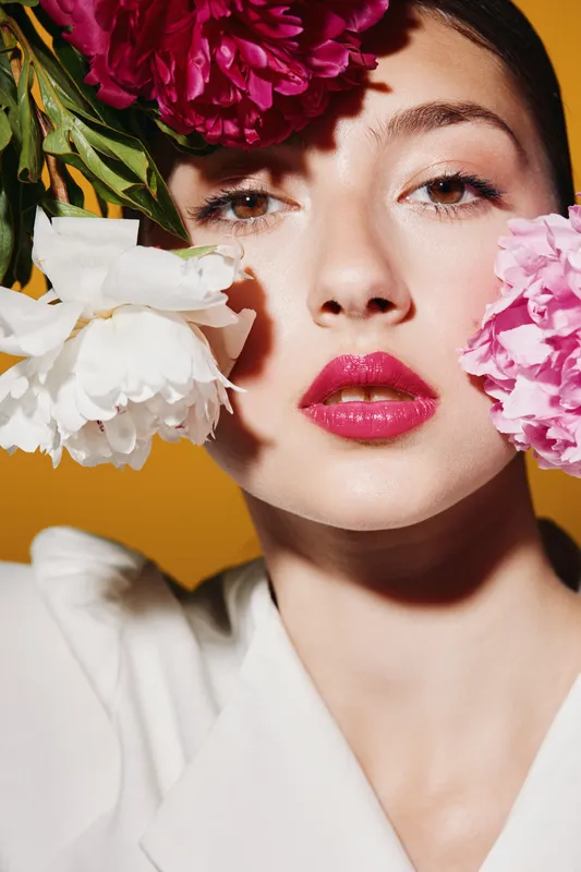 Close-up of fashion model with glossy pink lips surrounded by vibrant peony flowers on yellow background. Creative beauty shot emphasizing color and texture.