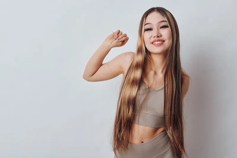 Young woman with long hair wearing a crop top and skirt standing against white background. Smiling female posing with raised arm and relaxed expression in studio setting.