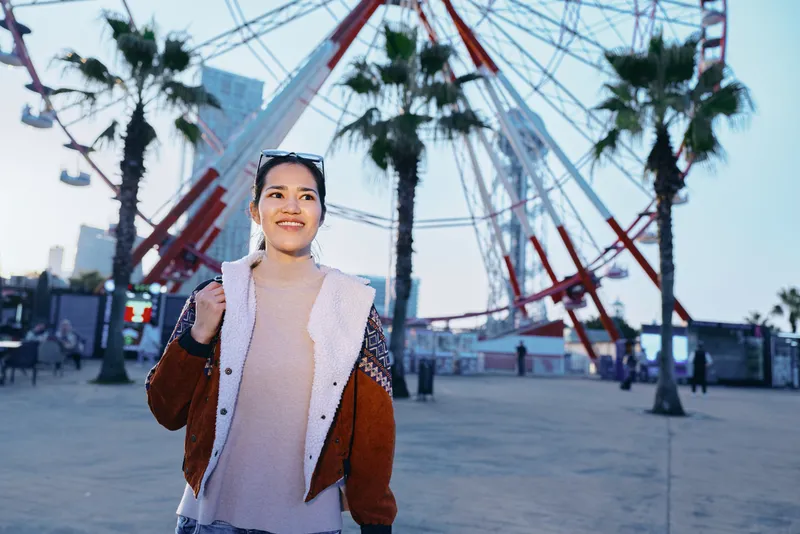 Smiling woman exploring an outdoor amusement plaza with a large ferris wheel, palm trees, evening light, casual jacket and sweater, embodying relaxed travel vibe and urban leisure in a photo friendly