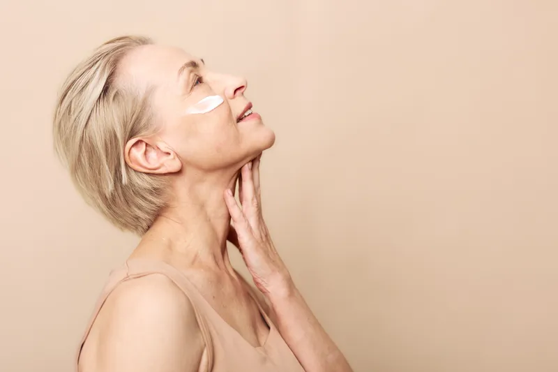 Middle aged woman with short blonde hair applies white face cream on cheek, eyes closed and hand on neck. Concept of skincare, aging care, beauty routine, and healthy skin glow.
