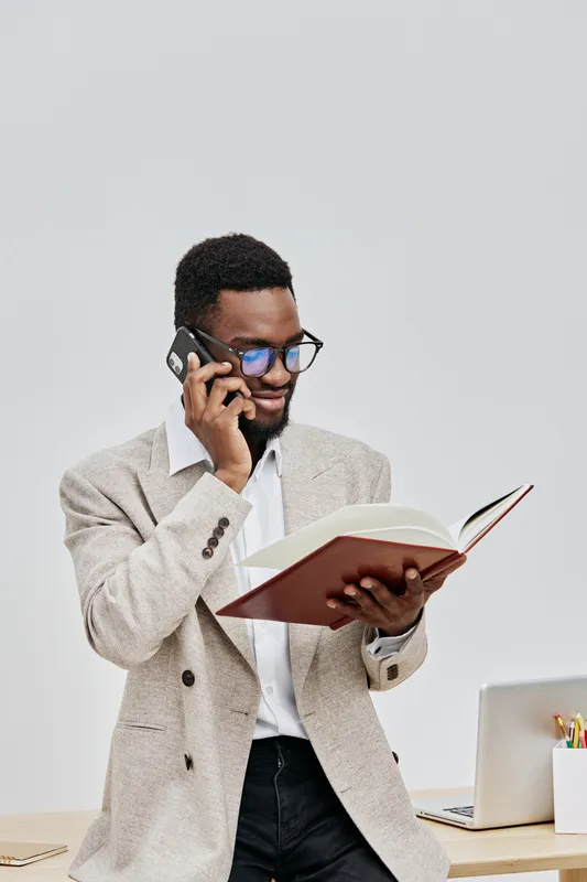 Confident young man with afro hairstyle and glasses reading a book while talking on mobile phone indoors with a plain white background, casual business outfit, professional lifestyle concept, modern c