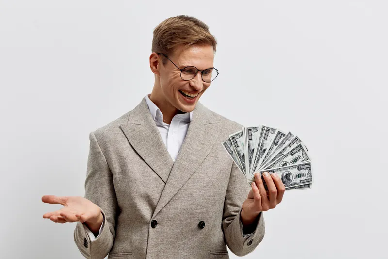 Confident smiling man in glasses holding fan of dollar bills in hand, dressed in stylish beige blazer, expressing success and wealth, isolated on plain white background, studio portrait, people lifest