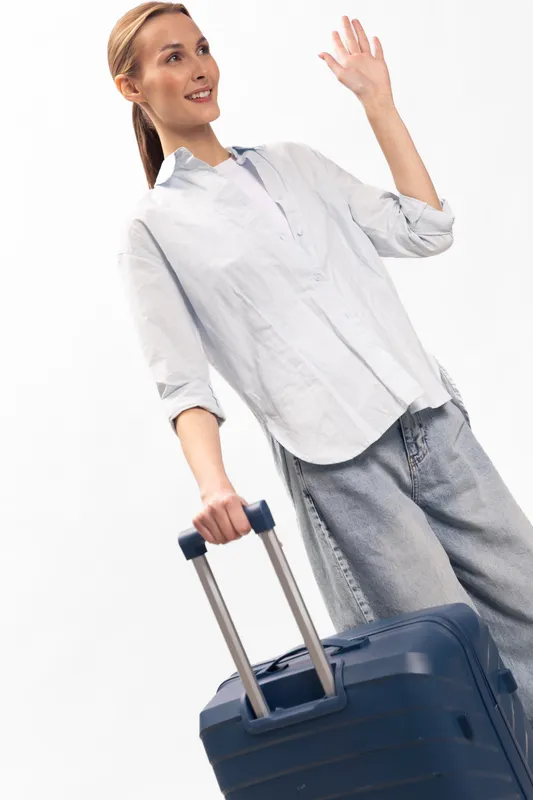 Young woman in casual clothes holding a suitcase with an extended handle waves goodbye against a white background. Concept of travel, departure, and vacation preparation.