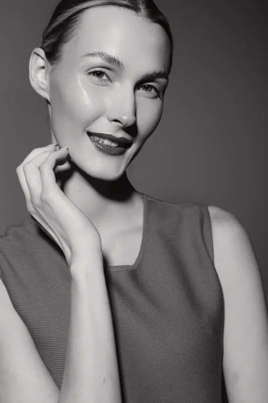 Smiling white woman with fair skin and slicked hair poses confidently in sleeveless top on dark background. Concept of beauty and natural glow.