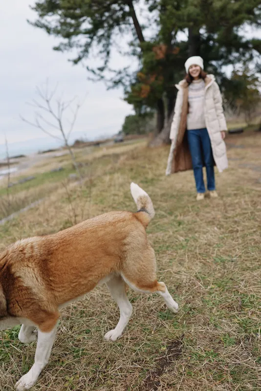 Woman walking dog across an open field in a warm coat, outdoor scene with trees in the background, capturing a calm lifestyle and canine companionship on a cool day.