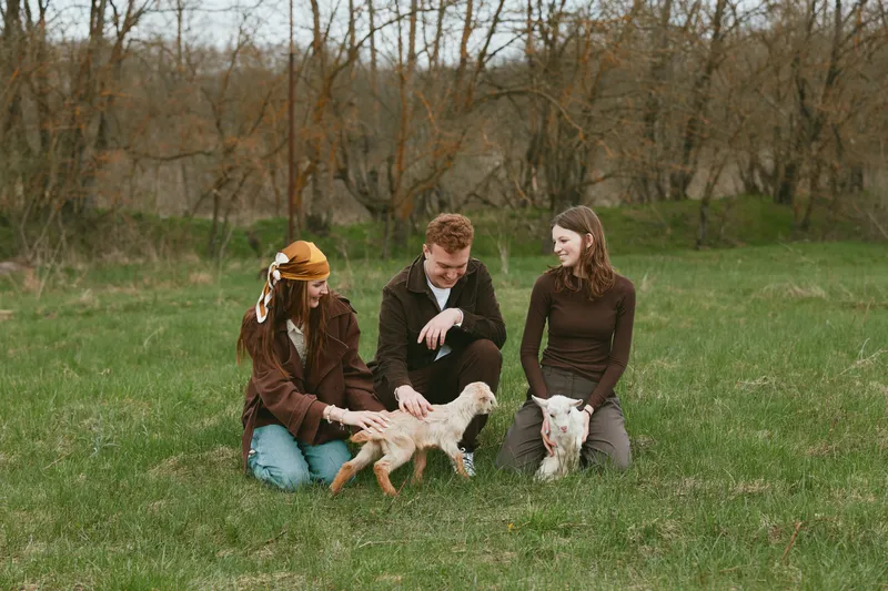 Three friends kneel in a grassy field, gently petting two young goats. A candid lifestyle moment focused on outdoor living, playful interaction, and calm connection with animals in a rural setting.