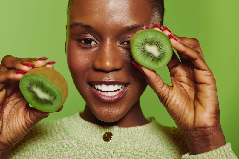 Smiling dark skinned African woman holds two halves of fresh kiwi fruit near face on green background. Concept of healthy lifestyle, natural food, wellness, and vibrant mood.