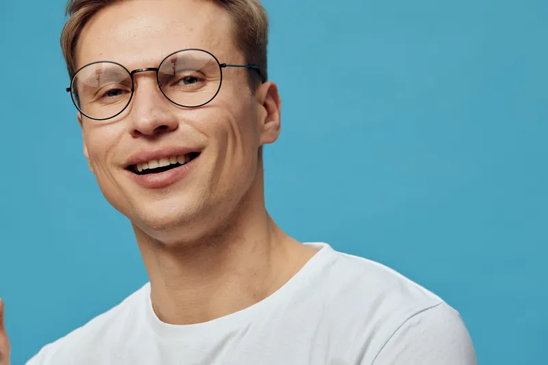 Smiling young Caucasian man with glasses wearing white T-shirt isolated on solid blue background, casual style, friendly expression, relaxed pose, cheerful mood, studio portrait. People lifestyle conc