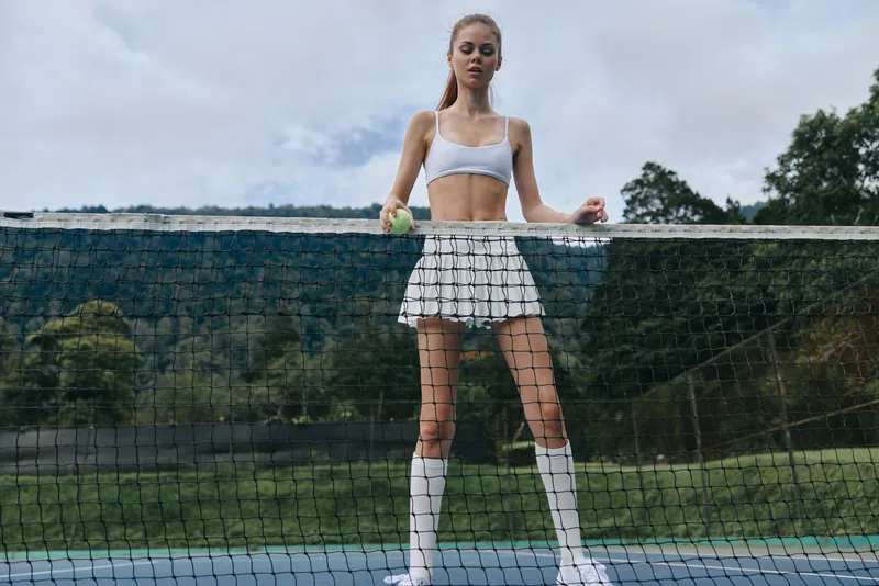 Young female tennis player in sporty attire, looking focused and ready to serve a tennis ball on a vibrant outdoor court surrounded by nature